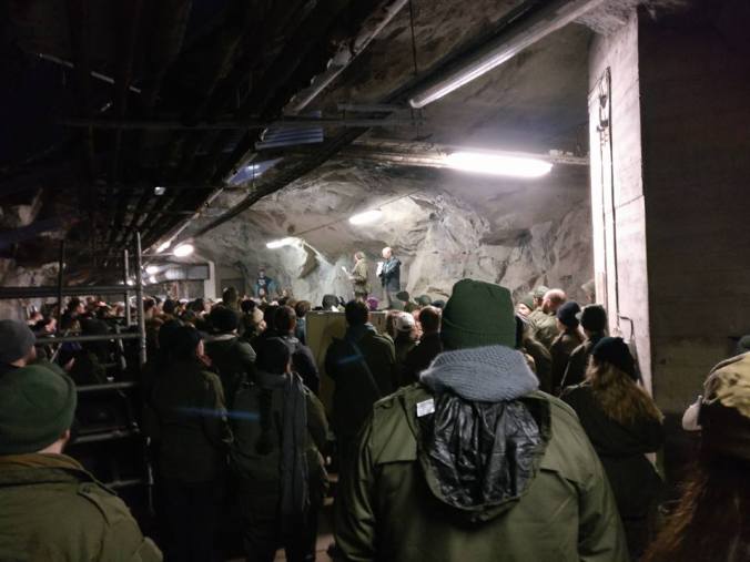 Ihmisiä bunkkerissa kuuntelemassa korokeella puhuvia henkilöitä, halogeenivaloa, armeijakuoseja ja värimaailmaa.