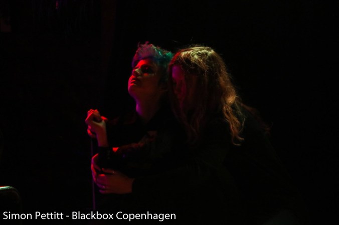 Kuvassa kaksi punaisen valon valaisemaa ihmistä syleilyssä mustassa ympäristössä.