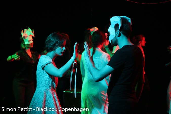 Kuvassa paperiseen naamioon pukeutunut ihminen kohottaa kättään ja naamioitta oleva ihminen kohottaa kättään kuin koskettaakseen tätä kättä. Taustalla lisää ihmisiä naamioissa.