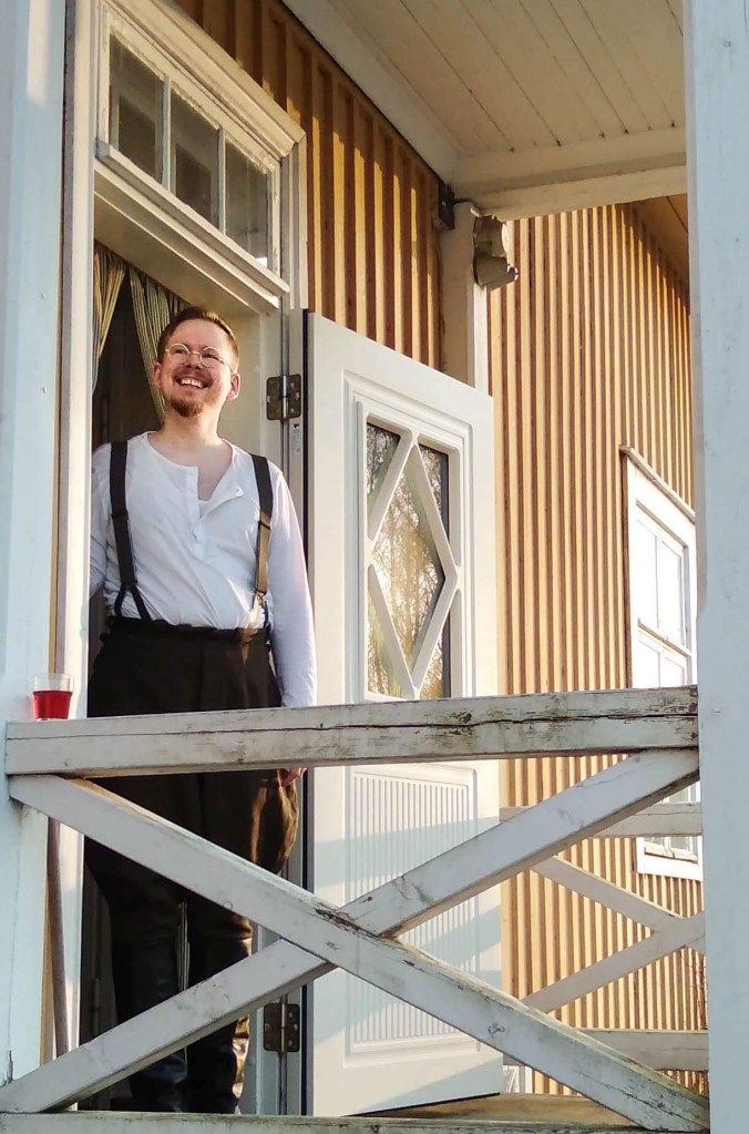 pelinjohtaja seisoo keltaisen puutalon ovella valkoisessa sarkapaidassa ja pussihousuissa