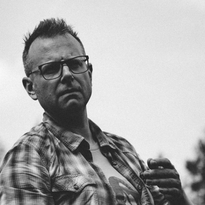 Mustavalkoisessa kuvassa Jon Hodgson näyttää vakavalta. Hänellä on lyhyt tukka, ruutupaita t-paidan päällä, ja hän näyttää selittävän jotakin.