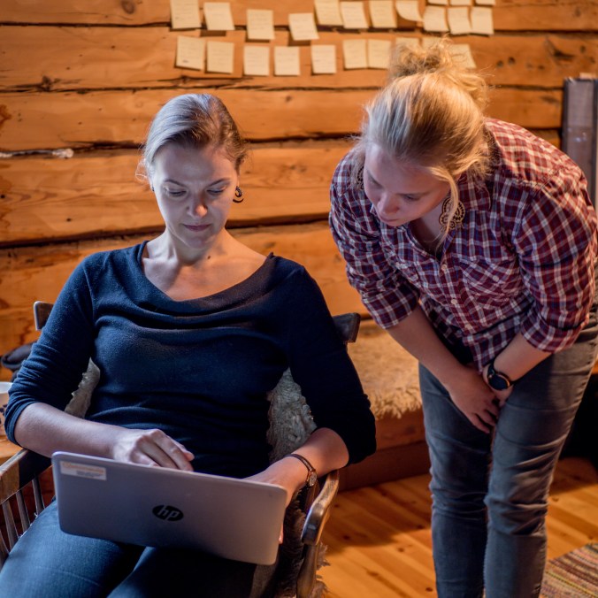 Kuvassa kaksi naista katsovat kannettavan tietokoneen ruutua, toinen istuu keinutuolissa, toinen seisoo, taustalla hirsiseinä, jossa post-it lappuja