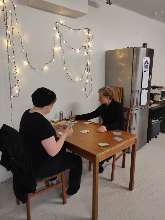 kaksi ihmistä tuolin pöydän ääressä pelaamassa korttia, taustalla pieni toimistokeittiö ja jouluvalot