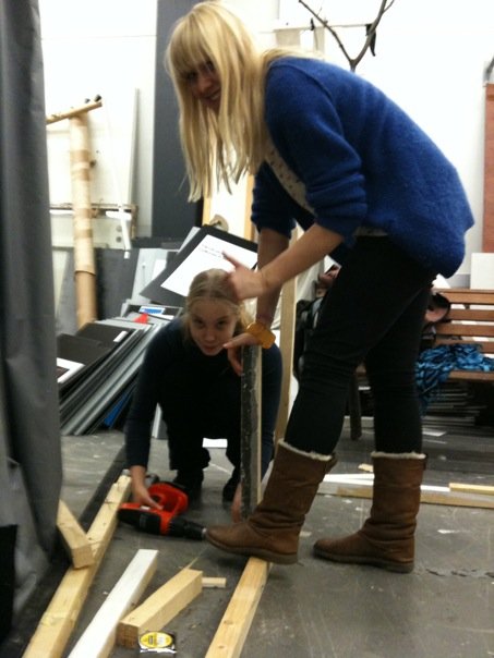 Kaksi nuorta naista poraavat lautoja yhteen lavasteita tehden.