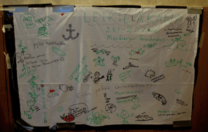 Kuvassa valkoinen lakana jota on koristeltu tusseilla piirrustuksin ja tesktein, esim lokkeja, ihmisiä, kalanruoto, uiva hirvi.