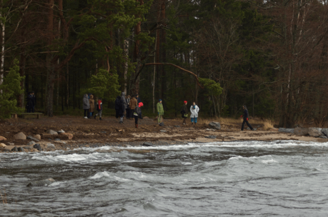 Kuvassa hahmoja meren rannalla syksyisen oloisessa tuulisessa säässä.