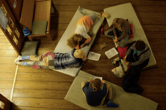 Kuvassa ihmiset istuvat lattialla patjojen päällä tekemässä tehtävää, joka ons elvästi annettu isoina paperinippuina joissa on paljon tekstiä.