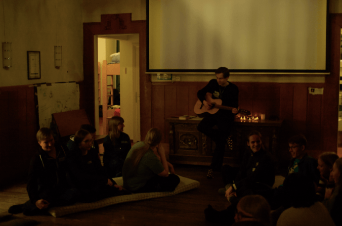 Kuvassa soitetaan kitaraa ja lauletaan, laulajat istuvat lattialla patjojen päällä. On hämärää, mutta kaapinpäällä on kynttilöitä.