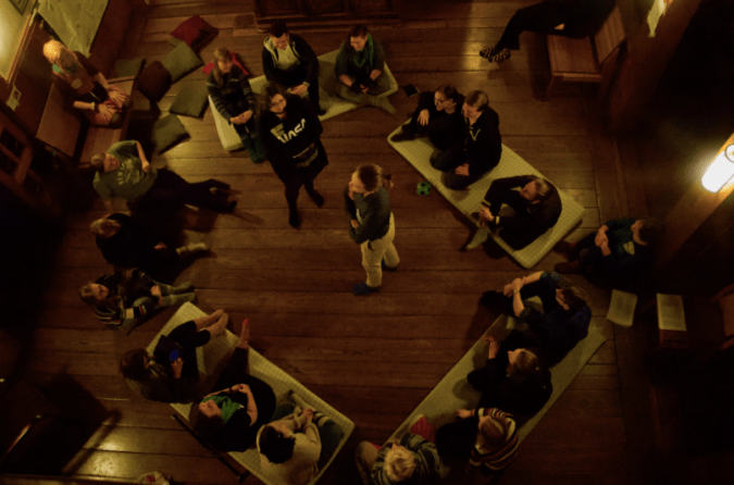 Kuvassa leikitään leikkiä, jossa muut istuvat ympyrässä lattialla ja kaksi "esittäjää" seisovat piirin keskellä.