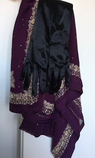 Kuvassa itämaisen näköisiä vaatteita roikkumassa naulassa, päällimmäisenä silkkiset mustat hanskat.