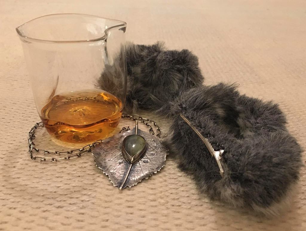 Kuvassa, harmaata turkista ja malja viskiltä näyttävää juomaa sekä hopeakoruja.