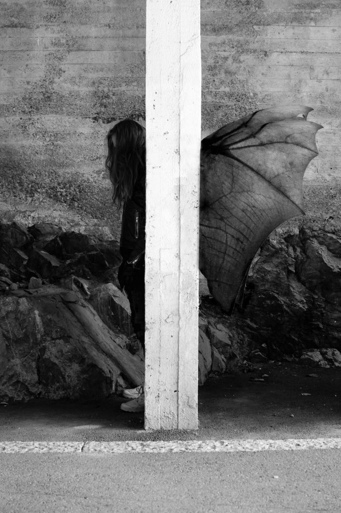 Asvaltilla betonipylvään takana seisoo sivuttain ihmishahmo pää vähän painuksissa. Pylvään toiselta puolelta näkyvät pitkät hiukset ja kädet ja toiselta puolelta harsomaiset siivet.