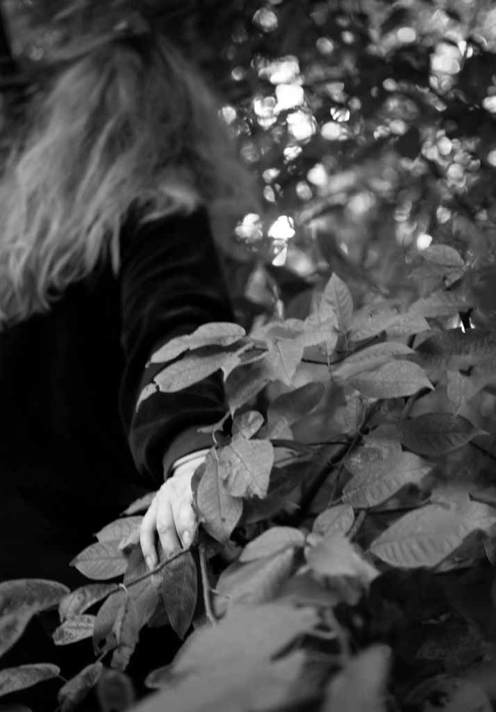 Pitkätukkainen ihmishahmo selin katsojaan siirtää syrjään oksia ja katoaa pensaikkoon.