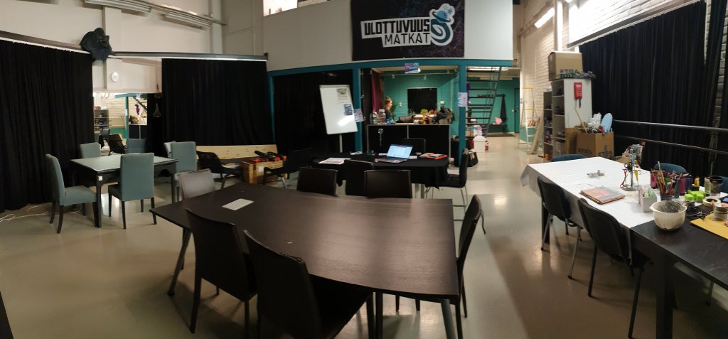 Kuvassa huone, jossa sienillä on akustiikkaa varten verhoja ja lattialla monta pöytää, joiden ympärillä on tuoleja. Seinällä lukee Ulottuvuusmatkat, pöydillä on erilaisia pelejä.