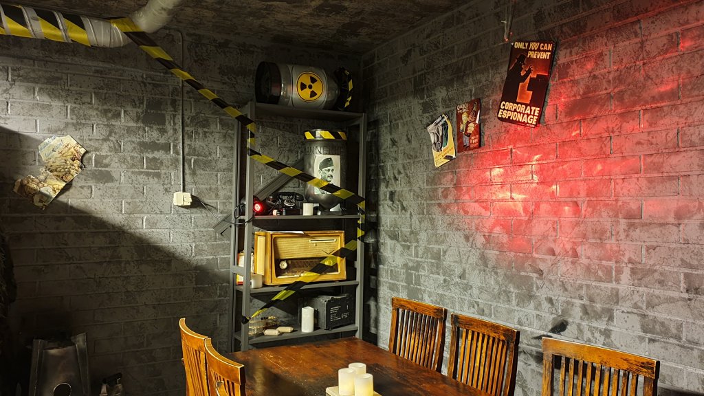 Kuvassa postapokalyptinen bunkkerihuone, eristysteippiä, harmaat tiilisienät, hyllyllä radiokaktiivisesta materiaaleista varoittavin tarroin koristeltuja esineitä. Pöytä ja tuolit.