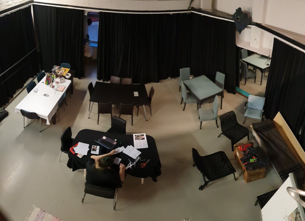 Sama pelitila kuin aiemmassa kuvassa, nyt ylhäältä päin kuvattuna: verhot sienillä akustiikkaa varten, pöytiä ja tuoleja pelaamiseen sopivina pieninä ryhminä.