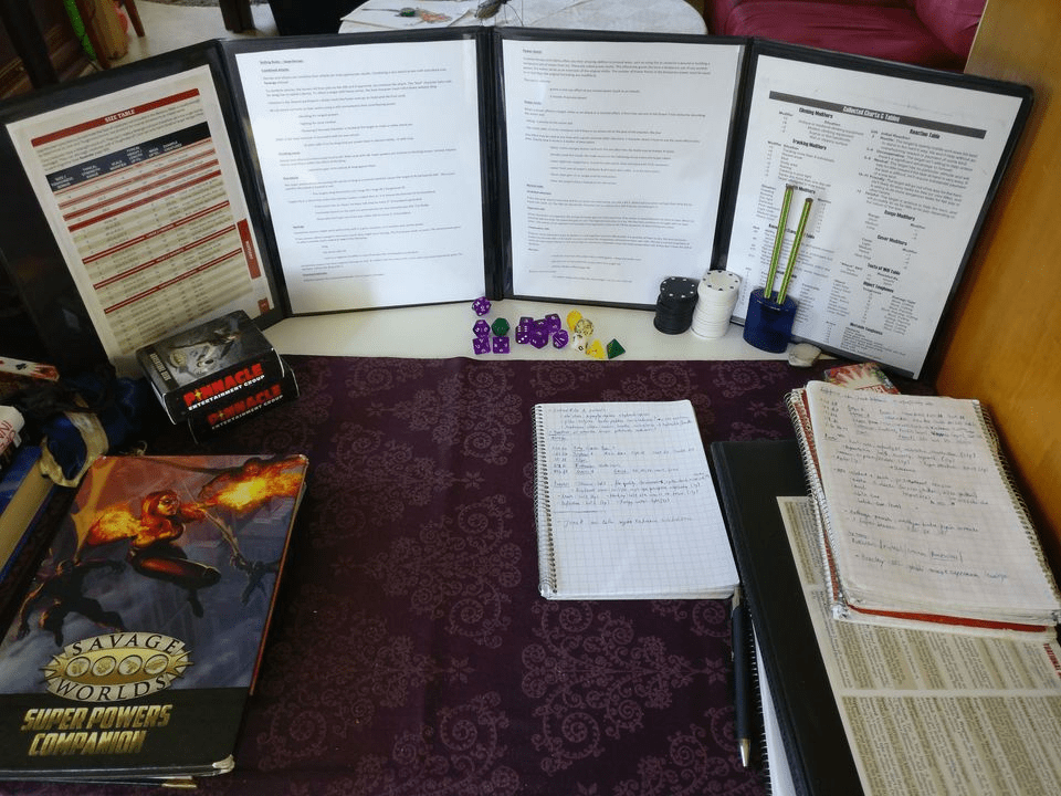 Kuvassa pelinjohtajan puoli pöytää näkösuojan takana, roolipelikirjoja, muistivihkoja, noppia, kortteja, kyniä.