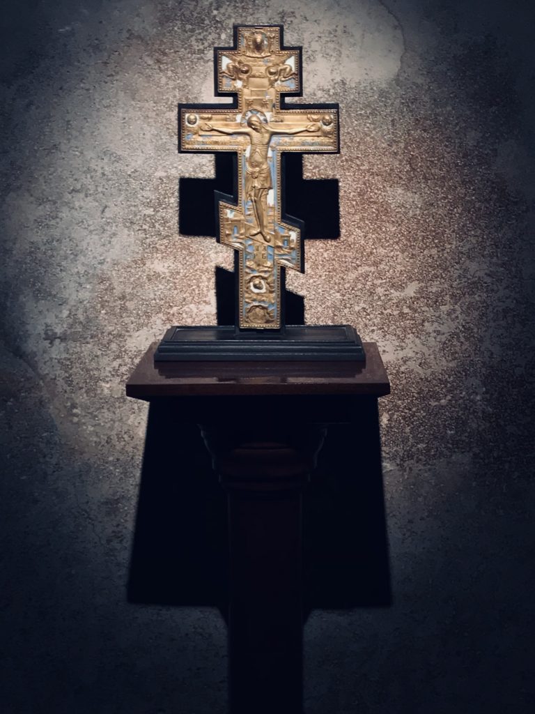 Kuvassa on kultainen ortodoksiristi, jossa on kuvattuna Jeesus ristillä. risti seisoo kiviseinää vasten puujalustalla ja siihen osuu kohdevalo.