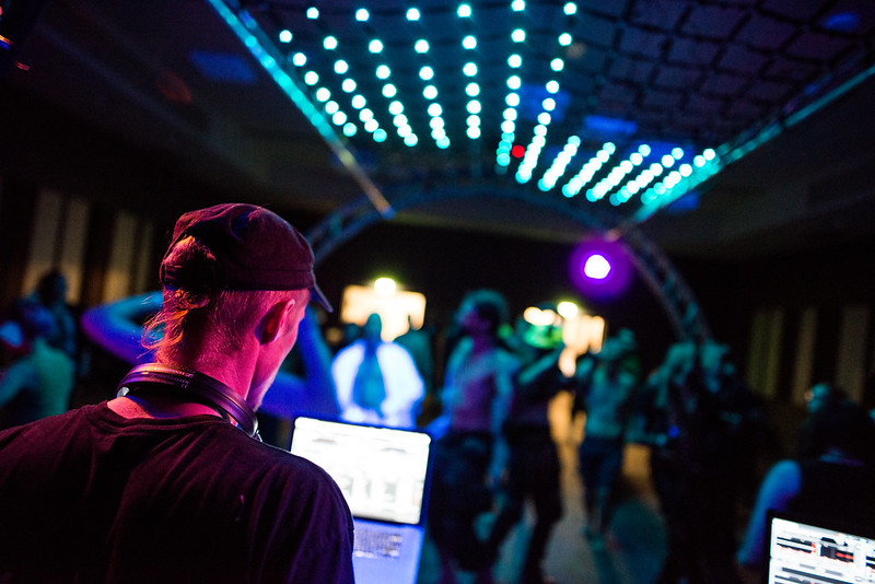 Kuvassa miesoletettu katselee tietokone-näyttöjä kuulokkeet roikkumassa kaulallaan selkä kameraan päin. Hänen edessään aukeaa tanssivien ihmisten sumuinen lauma diskovalojen valaisemana.
