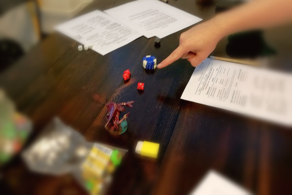 Kuvassa pöydällä roolipelilomakkeita, kumeja, figuureita, karkkeja, kyniä ja pyyhekumeja, joita ihmisen sormi osoittaa.